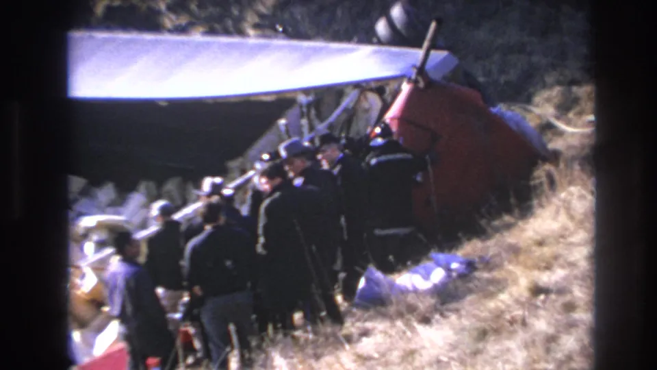 Hoboken 1966 8mm still showing men in dark clothing around red helicopter wreckage, with white debris and dry hillside grass nearby.