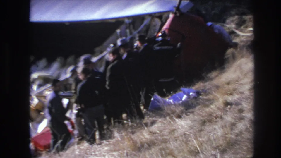Final 1966 Hoboken frame showing responders and suited men near overturned red aircraft wreckage on a dry grassy slope.