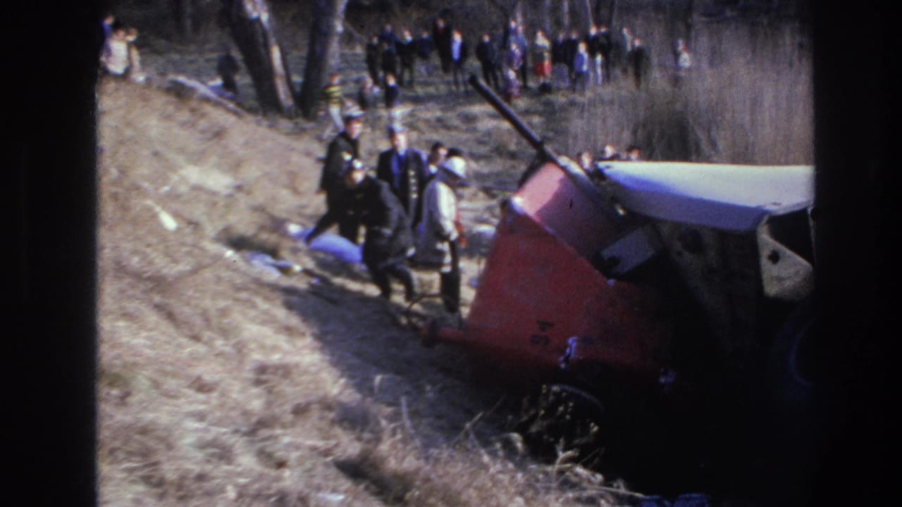 1966 Hoboken-area home-movie frame showing an overturned red-and-white truck beside a wooded slope with responders and onlookers.