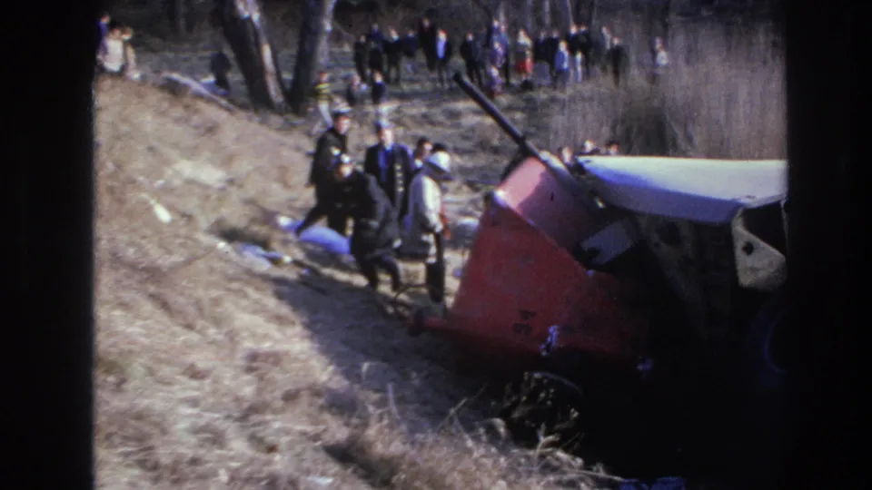 1966 Hoboken-area home-movie frame showing an overturned red-and-white truck beside a wooded slope with responders and onlookers.