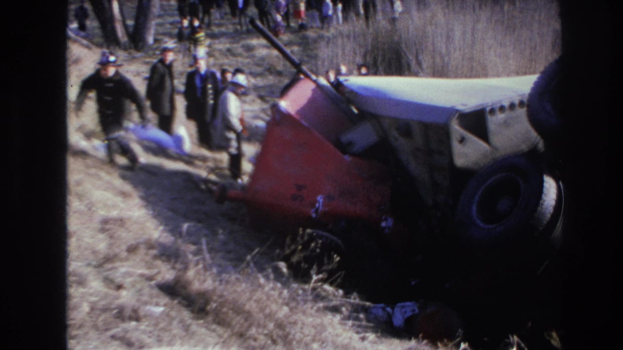 Responder and onlookers stand near the side of an overturned truck in a wooded ditch.