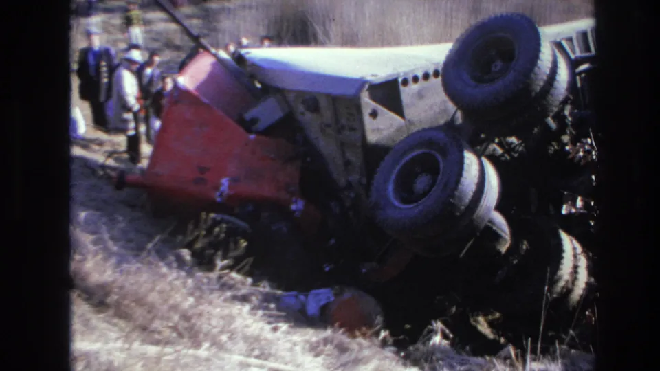 Closer 1966 home-movie frame showing the truck’s underside and wheels, with responders and bystanders partly visible beside the wreck.