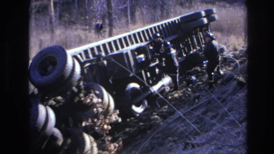 1966 Hoboken-area 8mm frame showing helmeted responders beside the overturned truck, with ropes or recovery lines crossing the foreground.