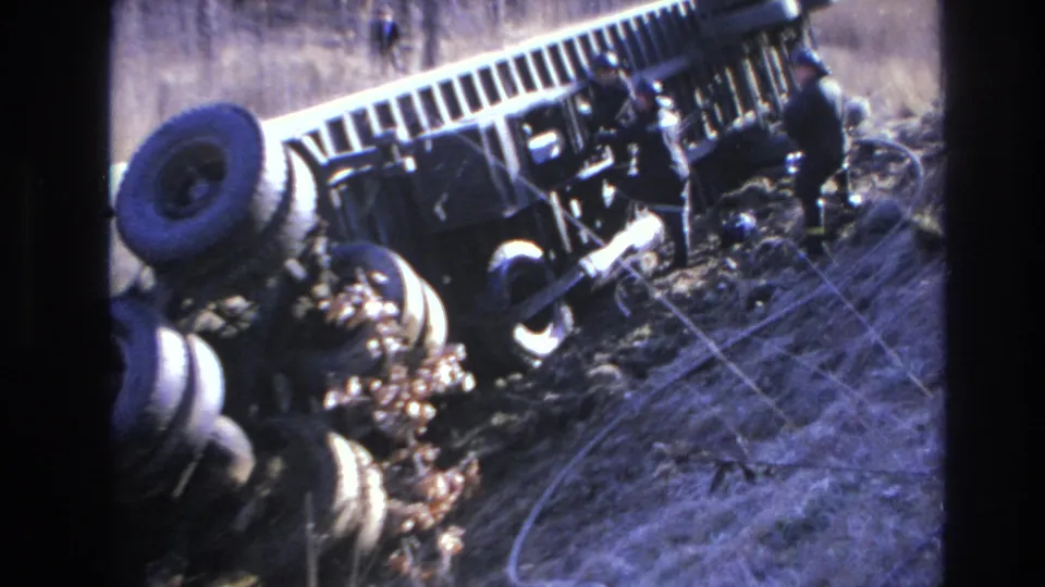 1966 Hoboken-area 8mm frame of the rollover aftermath, with responders near the truck and pale recovery lines across the dry grass.