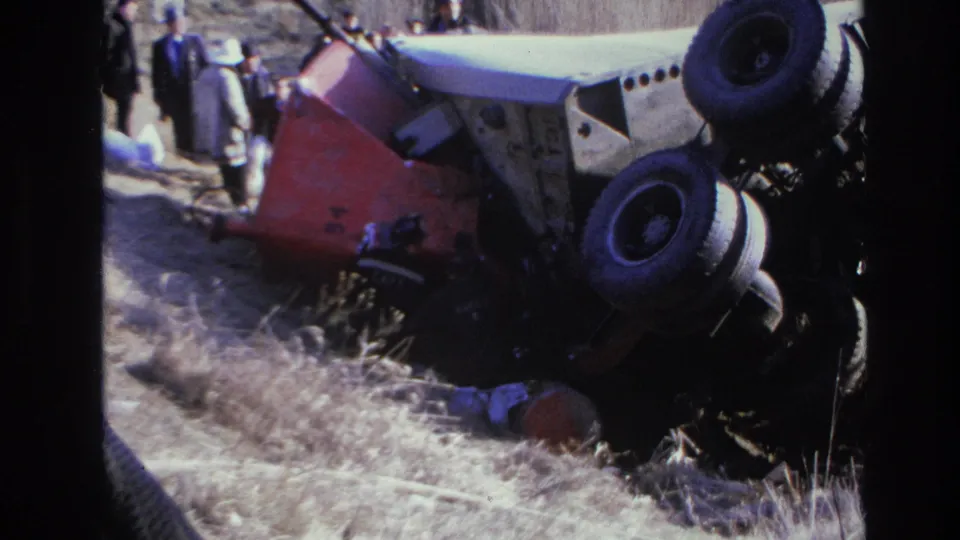 1966 Hoboken home-movie frame showing an overturned red and white car on a grassy embankment with people standing nearby.