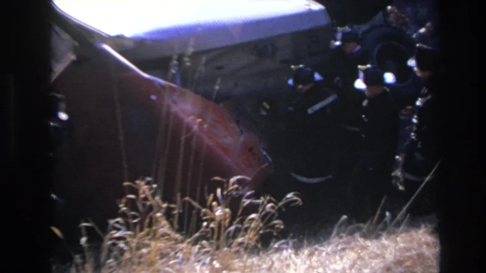 1966 Hoboken home-movie frame showing firefighters working near the overturned car, seen through soft 8mm focus and dark exposure.