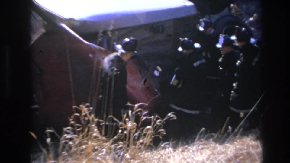 1966 Hoboken accident response frame with firefighters in helmets gathered around the overturned vehicle on a grassy roadside.