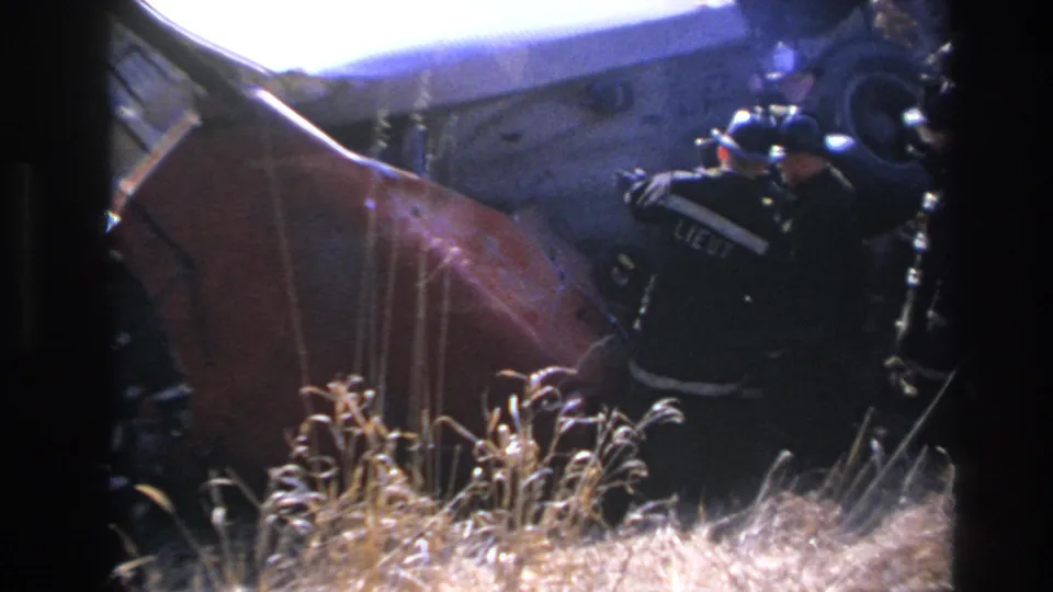 Nighttime 8mm frame from Hoboken showing a uniformed responder gesturing beside the overturned red vehicle.