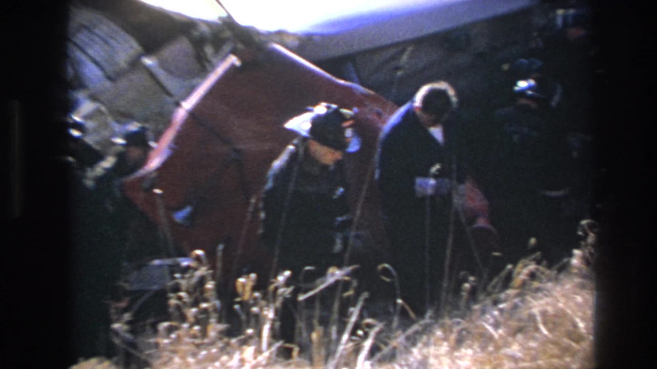 Uniformed responders in 1966 Hoboken appear beside a damaged red vehicle body, with dry grass and wreckage around them.