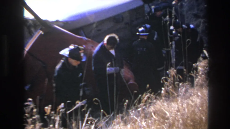Two uniformed figures stand near a damaged red vehicle body in the Hoboken crash-scene footage, partly obscured by dry grass.