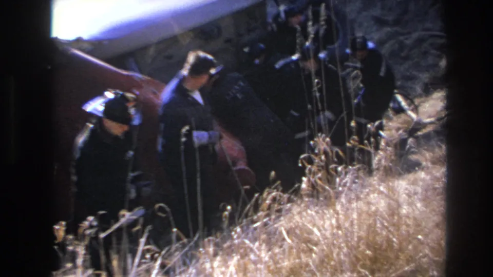 Final 1966 Hoboken frame shows responders beside the damaged red vehicle body, with one figure bending near wreckage and grass.