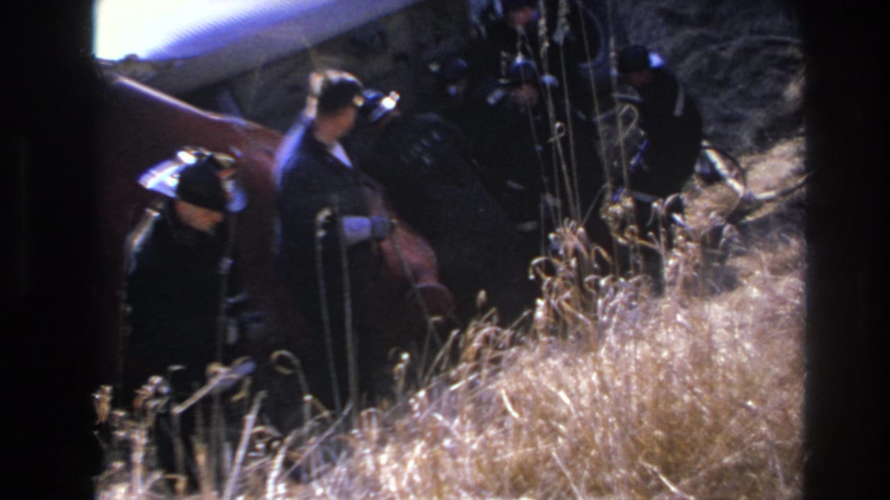 1966 Hoboken home-movie frame showing men in dark clothing and hats near tall dry grass, viewed through soft 8mm exposure and heavy frame borders.