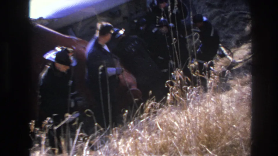 1966 Hoboken home-movie frame showing men in dark clothing and hats near tall dry grass, viewed through soft 8mm exposure and heavy frame borders.