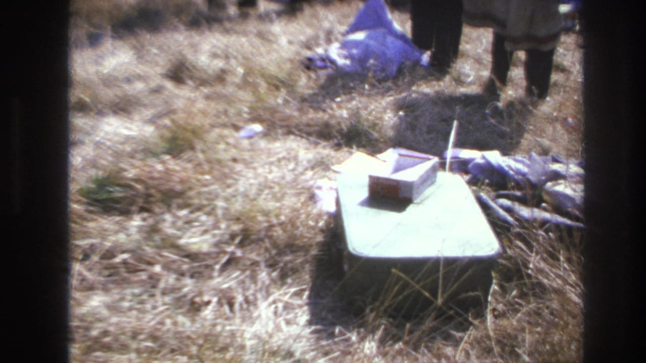 A green box or case sits in dry grass near scattered cloth and the lower legs of standing people.