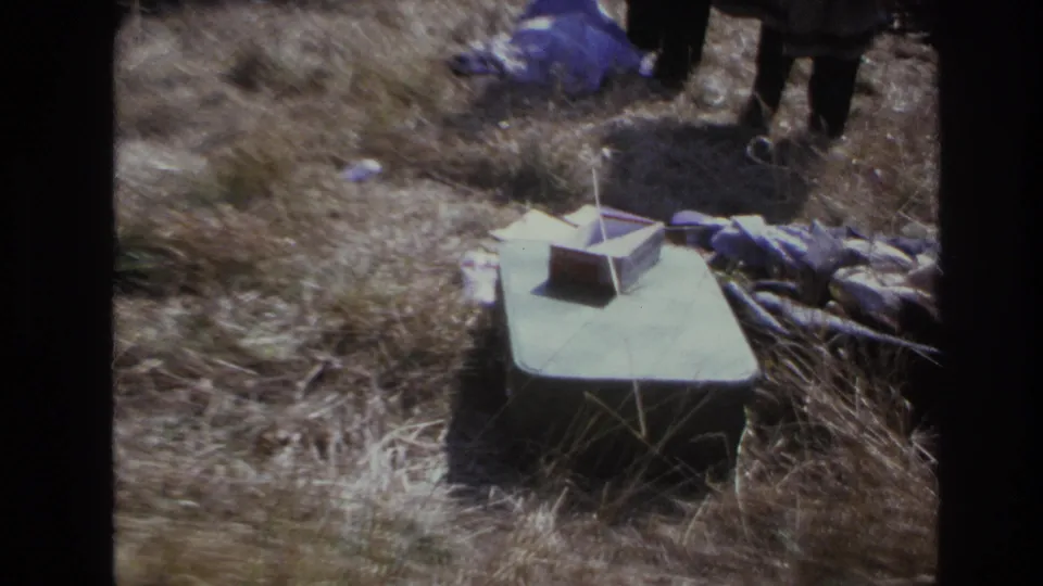 Closer 1966 Hoboken home-movie view of a green box in dry grass, a small object on top, fabric nearby, and standing figures at the edge.