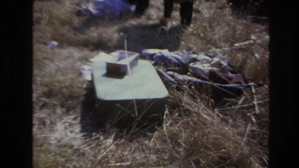 1966 Hoboken frame showing the green box, scattered fabric, dry grass, and standing figures in a soft, faded 8mm home-movie view.