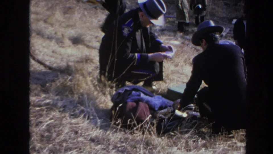 1966 Hoboken 8mm frame showing two hatted men crouched over discarded items in an overgrown field.