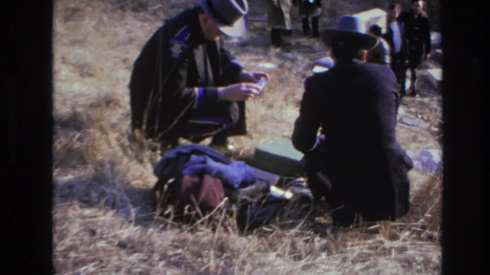 1966 Hoboken 8mm frame with two men in hats crouched over belongings as background figures watch from the field.