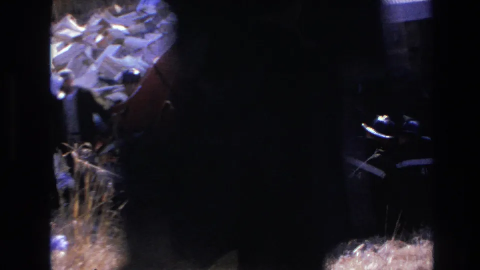 1966 Hoboken rescue scene with firefighters working beside an overturned red train car, seen through dark 8mm frame edges.