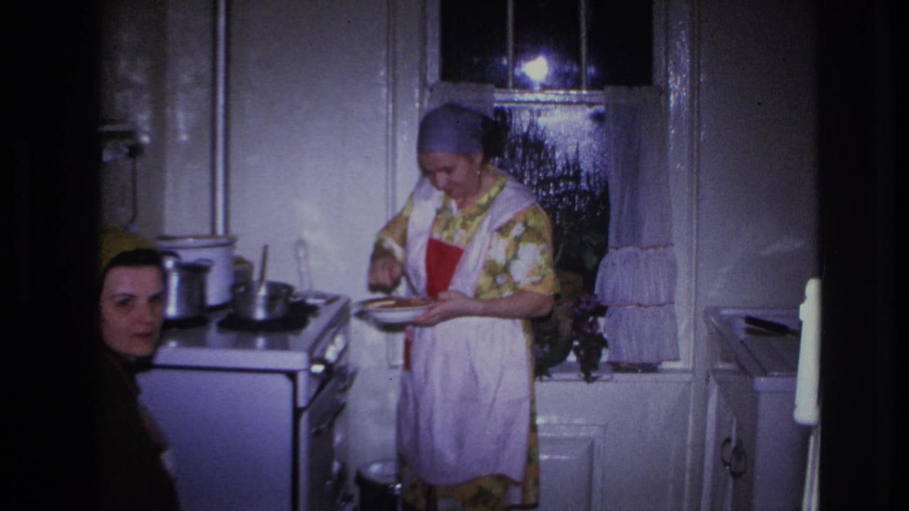 In a Hoboken 1966 home-movie frame, a woman in a floral dress and apron works over a plate beside a stove as another woman looks on.