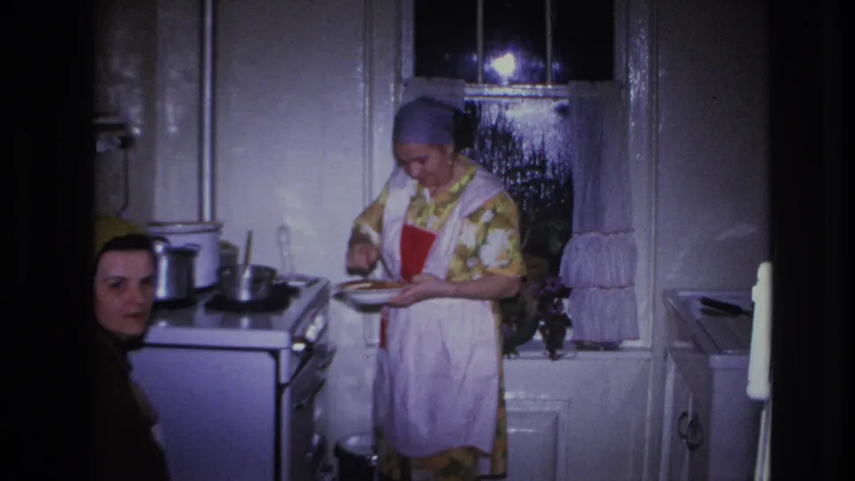 In a Hoboken 1966 home-movie frame, a woman in a floral dress and apron works over a plate beside a stove as another woman looks on.