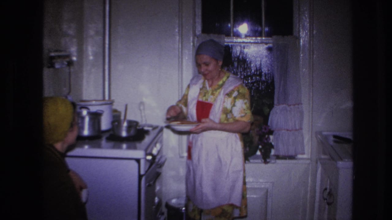 A woman in a gray headscarf and white apron prepares or serves food at a stove in a private Hoboken kitchen, with another woman partly visible.