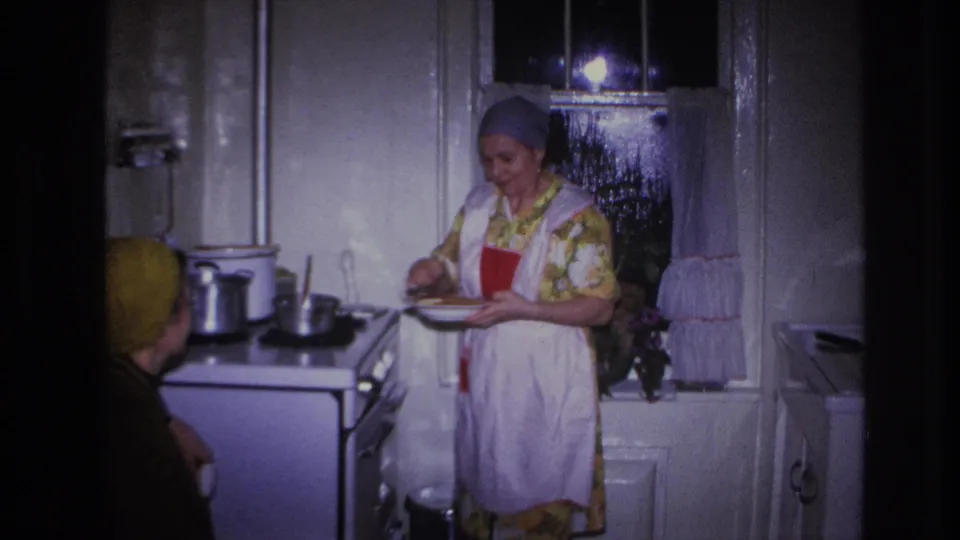 A woman in a gray headscarf and white apron prepares or serves food at a stove in a private Hoboken kitchen, with another woman partly visible.