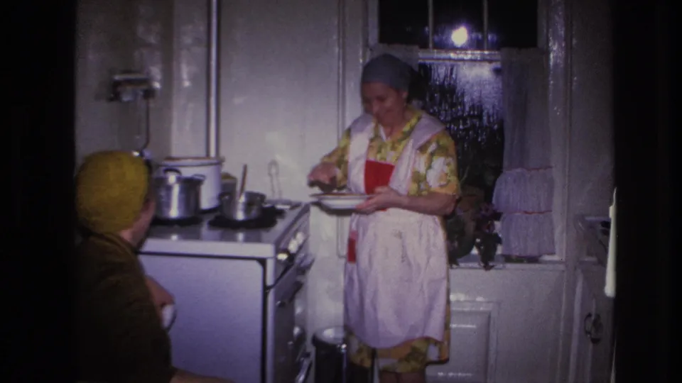 The seated woman watches from the foreground while the aproned woman continues preparing food near the stove in the 1966 Hoboken kitchen scene.