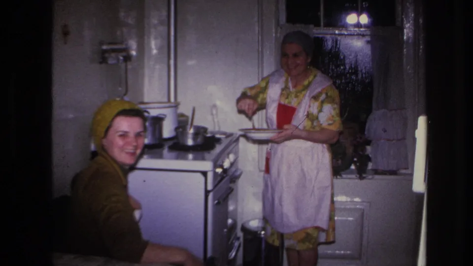 A smiling seated woman faces the camera while an aproned woman prepares food beside the stove in a private Hoboken home kitchen.