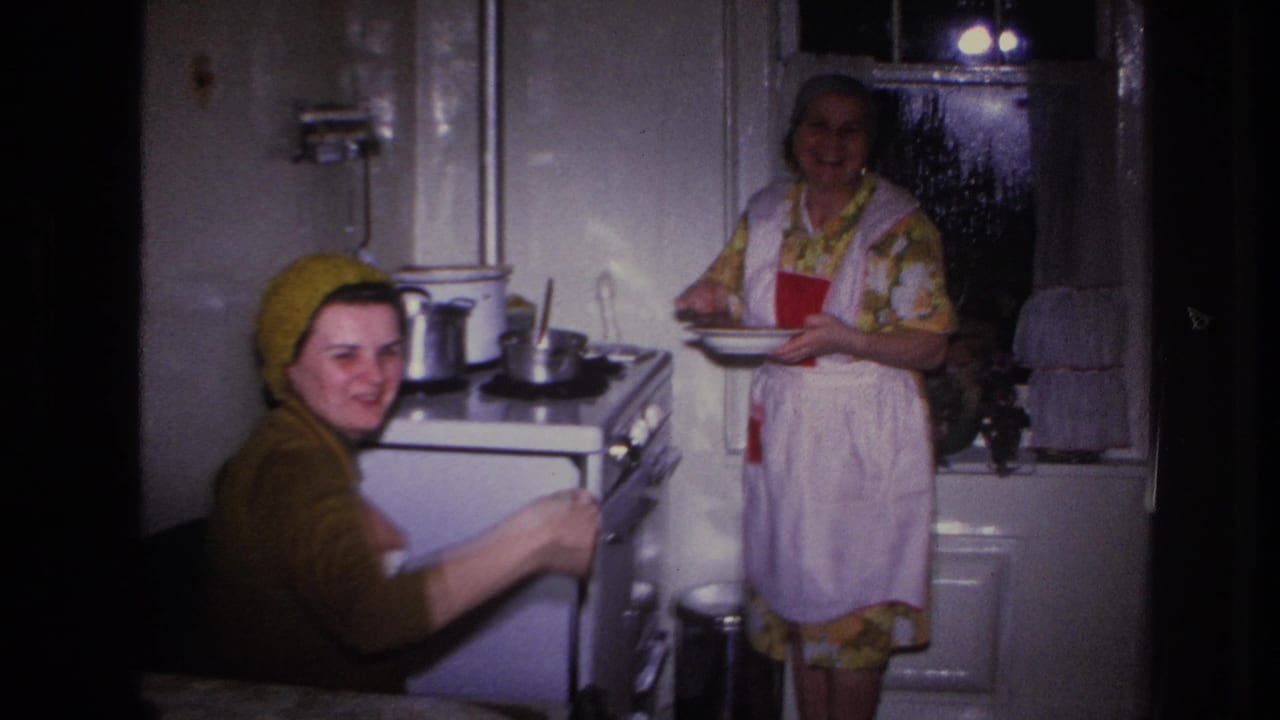 1966 Hoboken home-movie frame showing two women in a private kitchen with a stove, pots, apron, headscarves and curtained window.