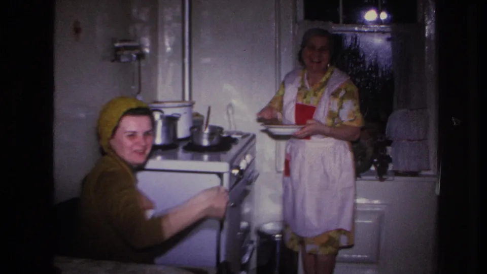 1966 Hoboken home-movie frame showing two women in a private kitchen with a stove, pots, apron, headscarves and curtained window.