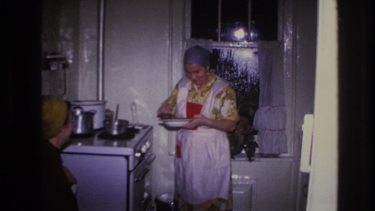 A woman in an apron holds a plate near the stove while another woman is partly visible in the foreground.