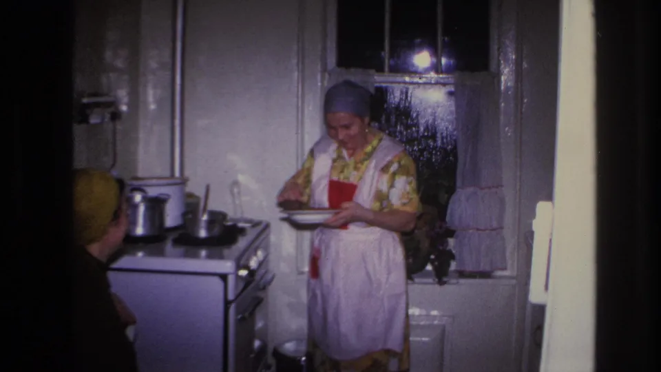 1966 Hoboken kitchen frame with an aproned woman looking down at a plate beside pots on a stove and another woman seated nearby.
