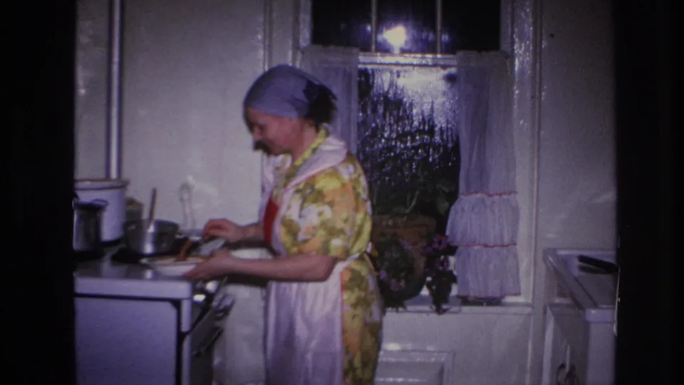 1966 Hoboken home kitchen frame showing a woman in a floral dress, headscarf and apron preparing food at a white stove.