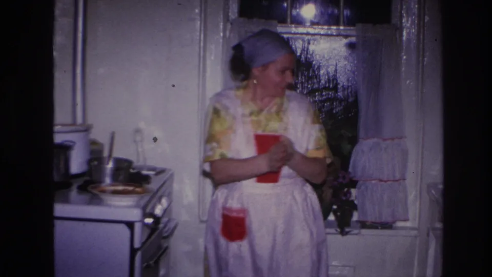 1966 Hoboken kitchen frame showing an aproned woman smiling near a stove, with cookware, curtains and a dark window behind her.