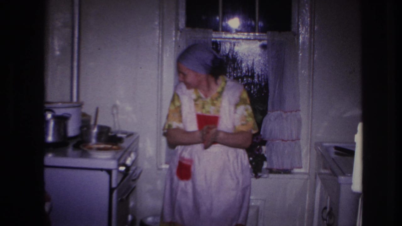 1966 Hoboken home-movie frame showing a woman in a yellow floral dress, white apron, and blue headscarf beside a stove in a dim kitchen.