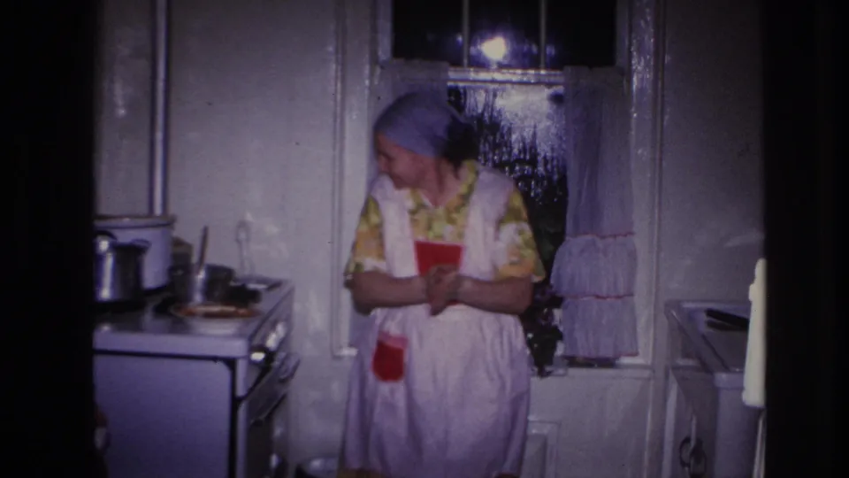 1966 Hoboken home-movie frame showing a woman in a yellow floral dress, white apron, and blue headscarf beside a stove in a dim kitchen.