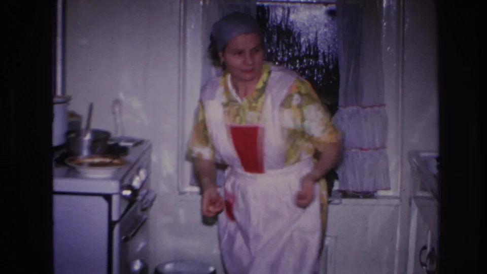 The woman turns in the small kitchen, wearing a yellow floral dress, white apron, and blue headscarf in the Hoboken home-movie scene.