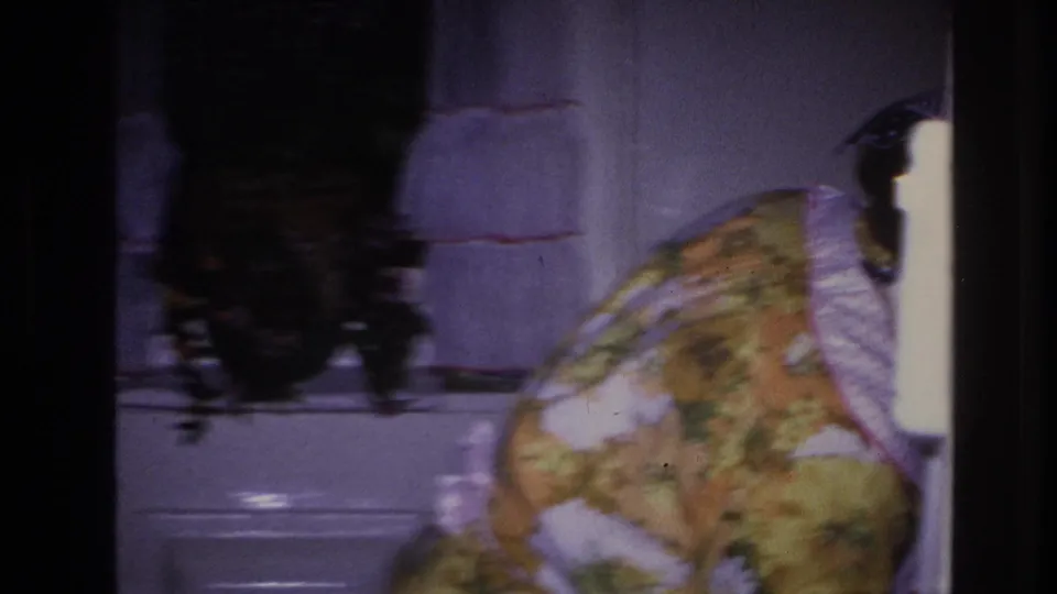 The woman bends near the right side of the compact kitchen, partly out of frame, with ruffled curtains and a sink or counter nearby.