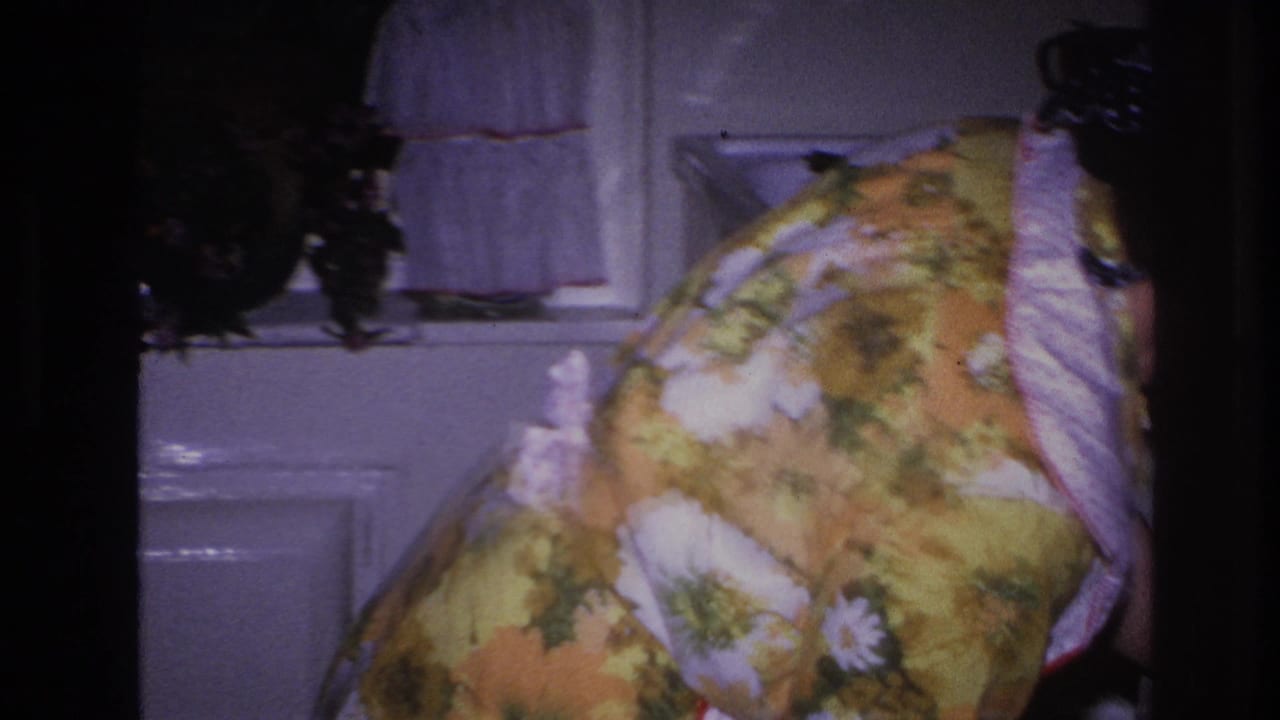 In a 1966 Hoboken home-movie frame, a woman in a yellow floral dress and apron bends near paneled doors inside a kitchen.