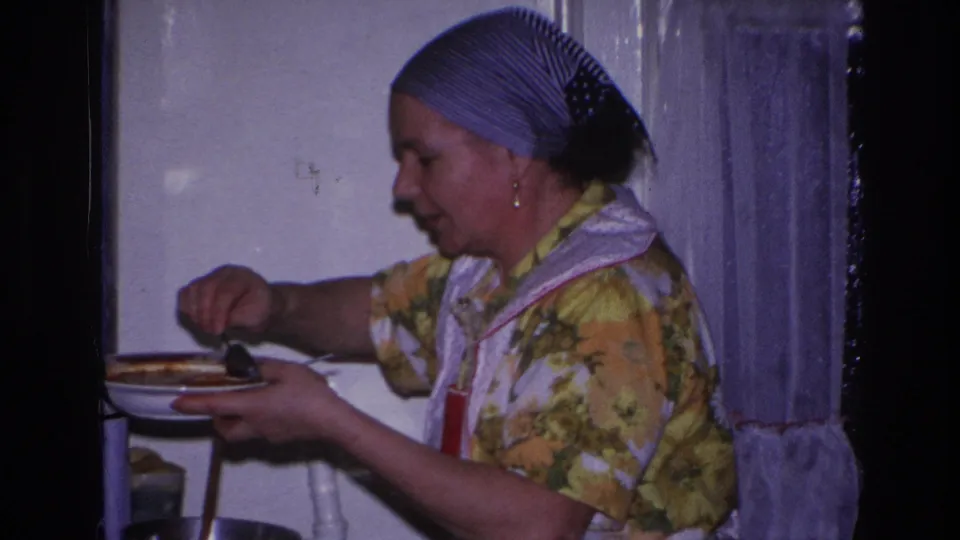The woman holds a plate and uses a utensil beside the stovetop, her headscarf and apron visible in the Hoboken home interior.