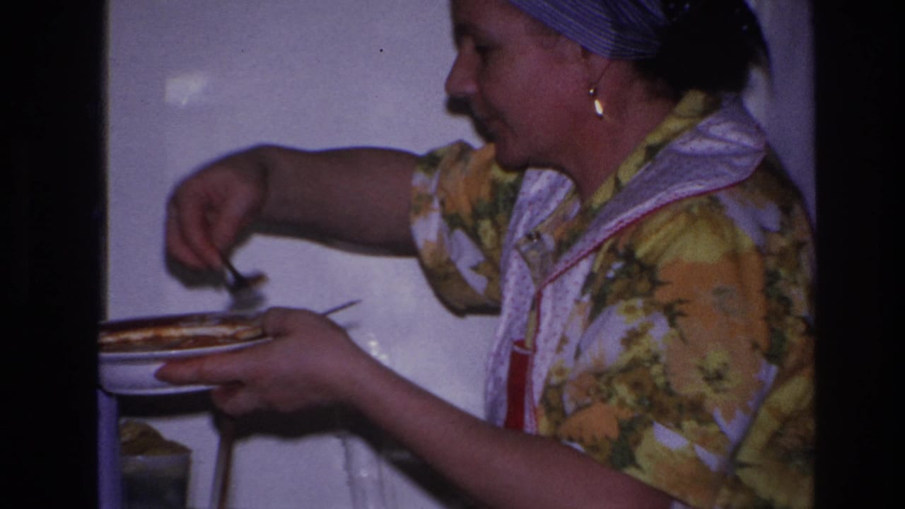 In 1966 Hoboken home-movie footage, a woman in a floral dress, apron, and headscarf serves food at a kitchen stove.