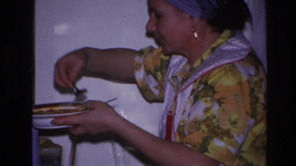 In 1966 Hoboken home-movie footage, a woman in a floral dress, apron, and headscarf serves food at a kitchen stove.