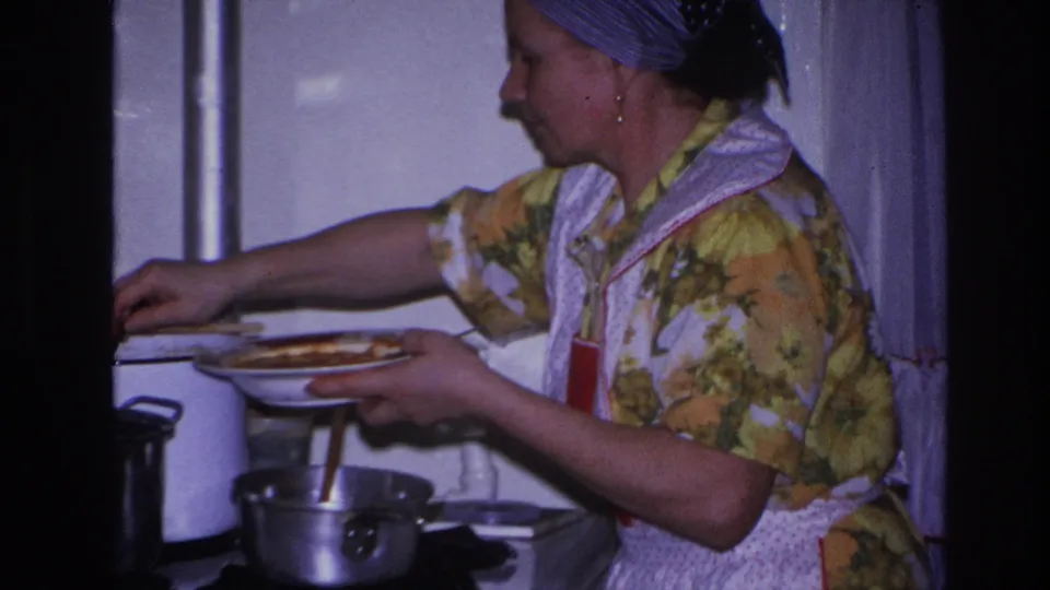 The woman reaches toward the stove area while holding a plate, with pots and curtains visible in the 1966 kitchen scene.