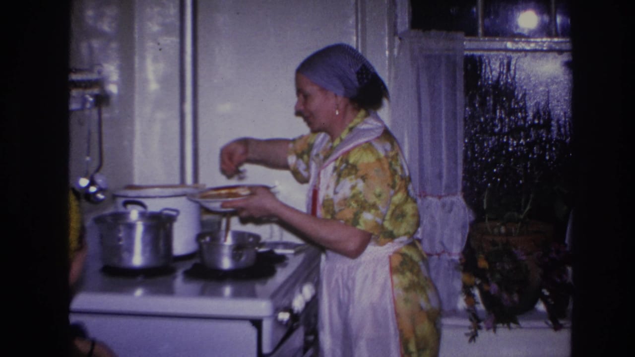 In 1966 Hoboken home-movie footage, a woman in a floral dress, white apron, and blue headscarf cooks at a white stove.