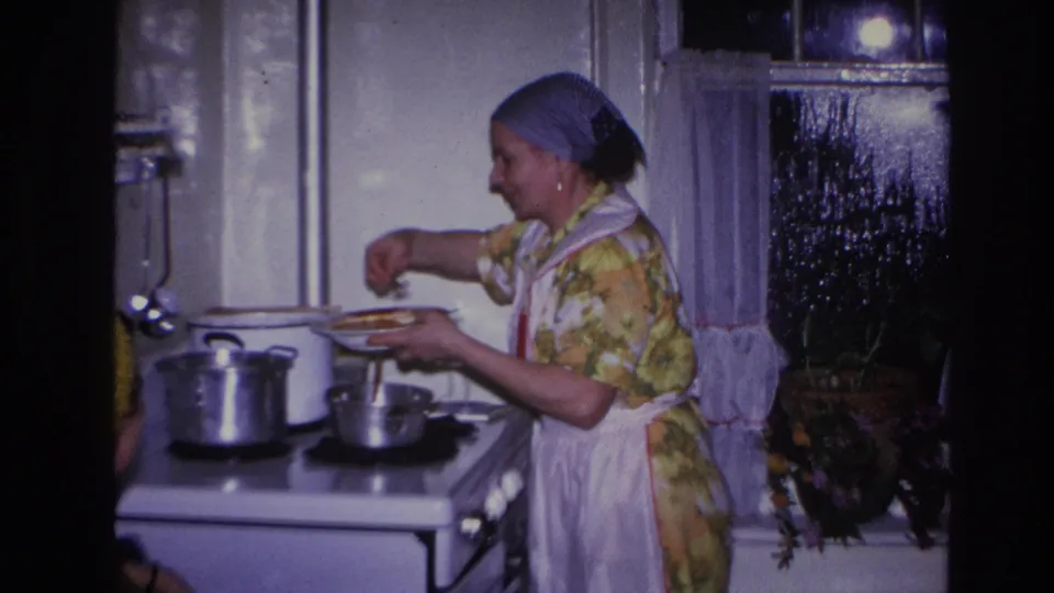 In 1966 Hoboken home-movie footage, a woman in a floral dress, white apron, and blue headscarf cooks at a white stove.