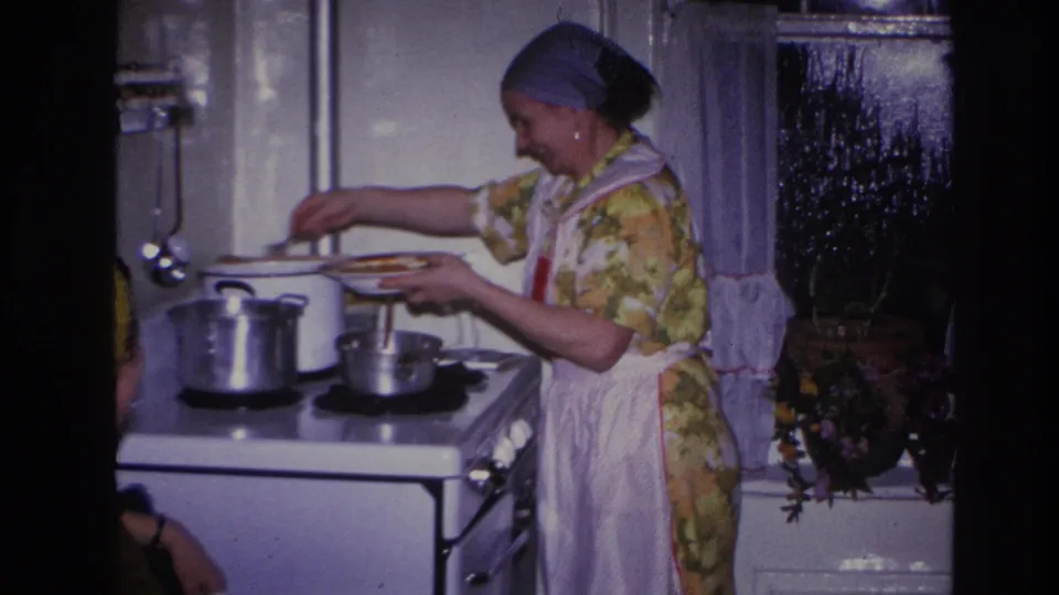 The woman stands beside metal pots on a white stove, holding a dish as she serves food in a 1960s Hoboken kitchen interior.