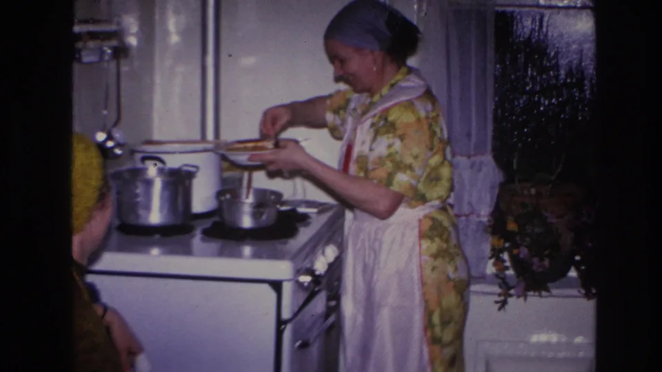 A woman continues serving food at the stove while a younger woman in a yellow knitted hat is partly visible in the foreground.