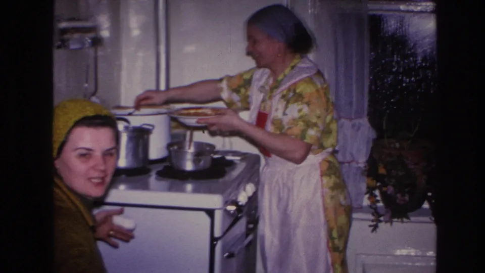 A younger woman in a yellow knitted hat looks toward the camera in the foreground as the cook smiles and serves food behind her.