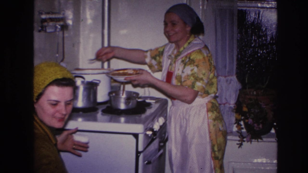 1966 Hoboken home-movie frame showing two women in a kitchen, with one serving food at a white stove and another smiling in a knit hat.
